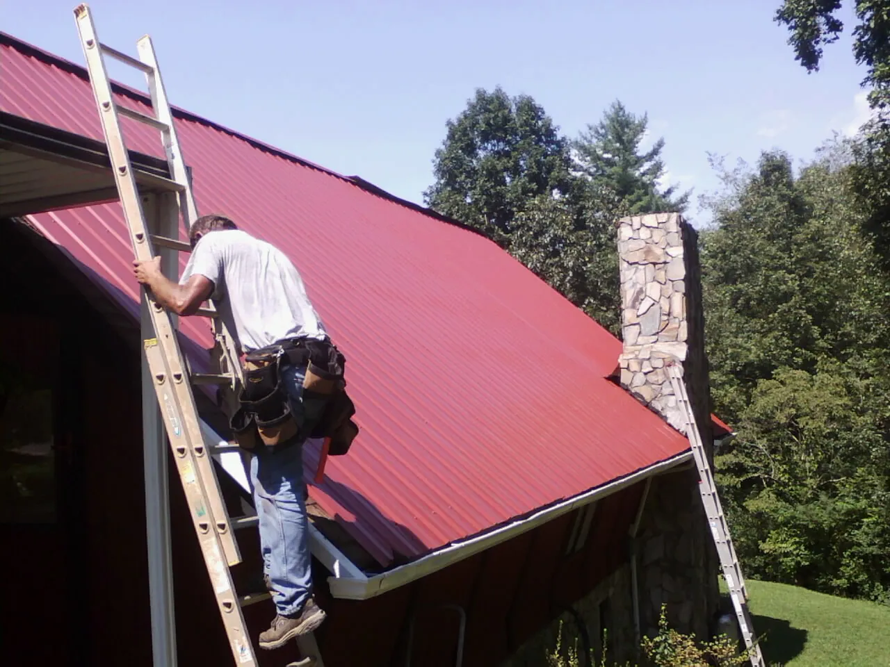 Roofer on a ladder beside a red metal roof and stone chimney — Soffit & Fascia Repair by Roofing Brothers in Dysart